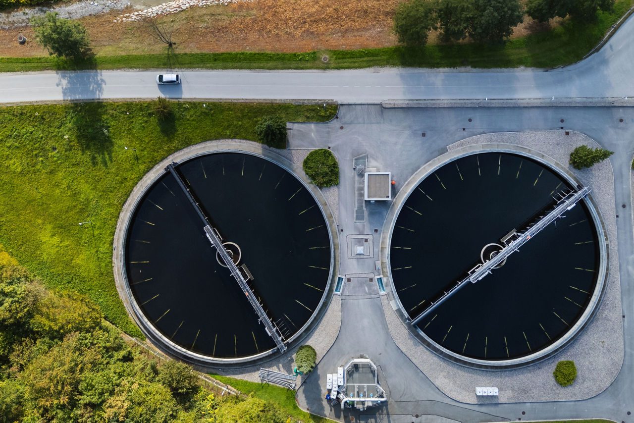 Vista dall'alto di un moderno impianto di trattamento delle acque reflue che mostra vasche di decantazione circolari e paesaggio verde integrato per la gestione delle acque.