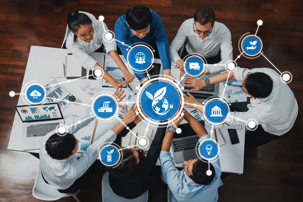 team eterogeneo riunito a un tavolo di lavoro e focalizzato sulla sostenibilità e l'innovazione, con le mani che si estendono su tutti i notebook e il simbolo del mondo per l'impatto globale.