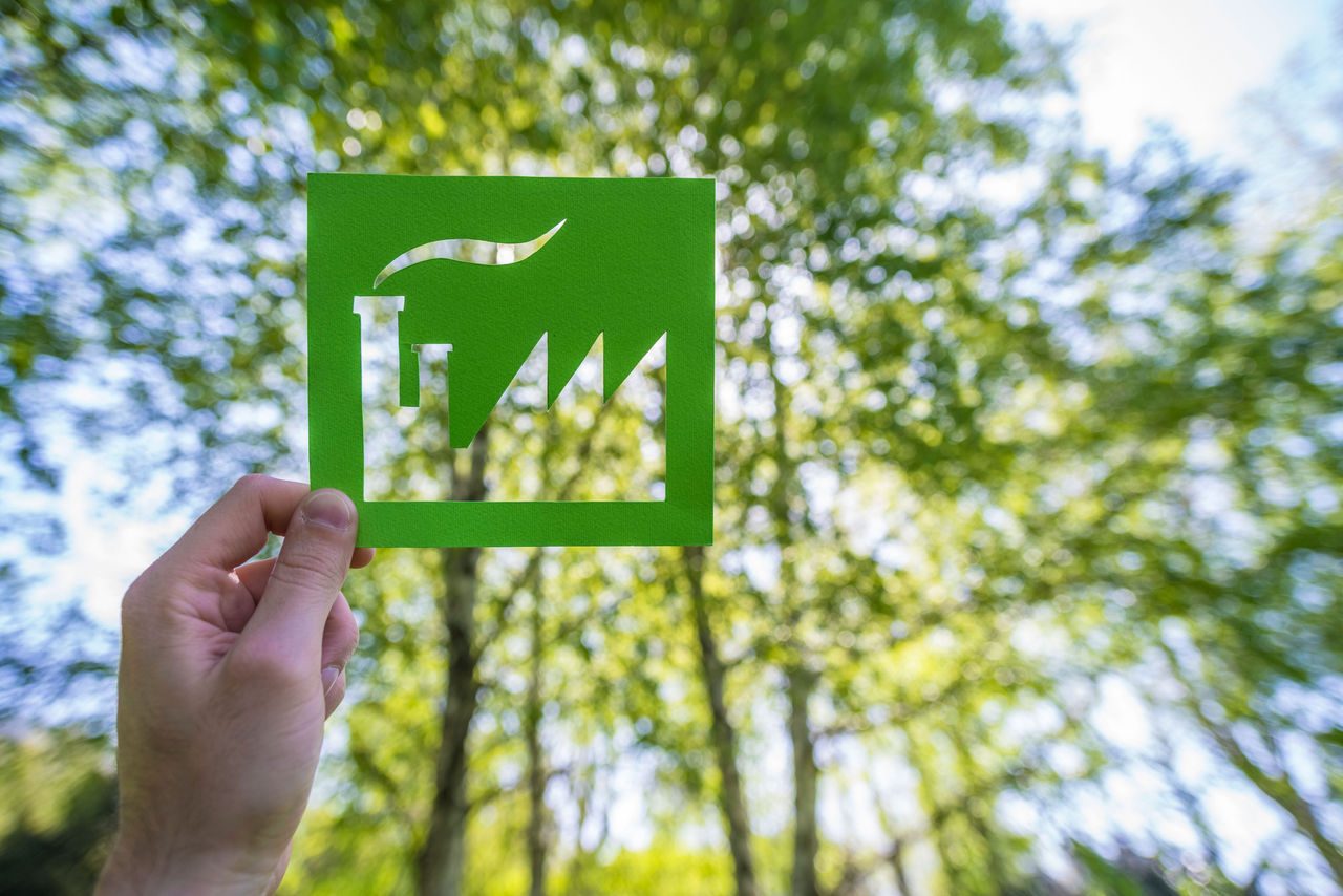 Una mano tiene un ritaglio di carta verde a forma di fabbrica con camino, simbolo della sostenibilità industriale, sullo sfondo di un bosco con alberi verdi e luce naturale filtrata tra le foglie. Una mano tiene un ritaglio di carta verde a forma di fabbrica con camino, simbolo della sostenibilità industriale, sullo sfondo di un bosco con alberi verdi e luce naturale filtrata tra le foglie.