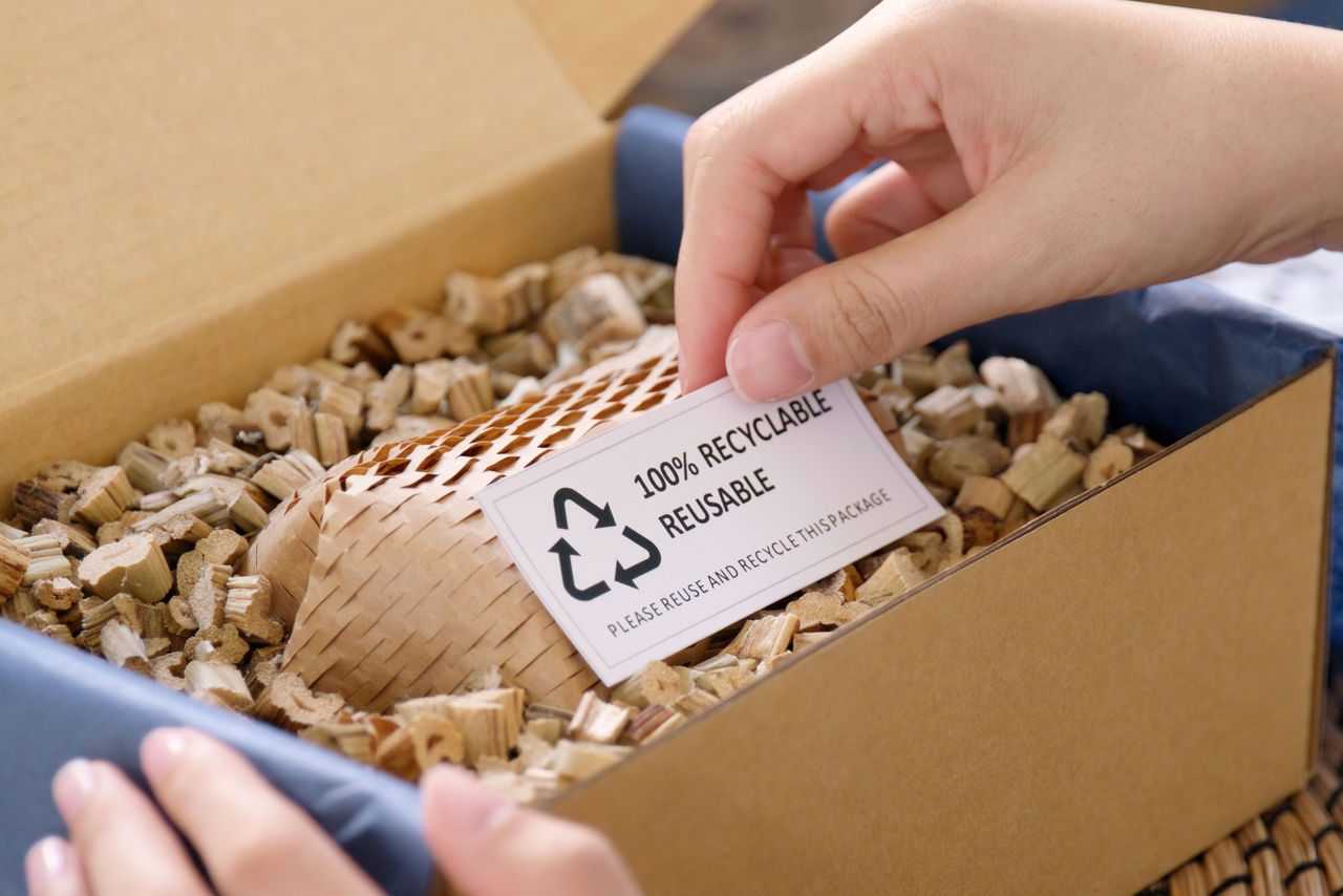 Una mano inserisce un'etichetta con la scritta "100% recyclable reusable" in una scatola contenente materiale da imballaggio ecologico, rappresentando il concetto di packaging sostenibile.