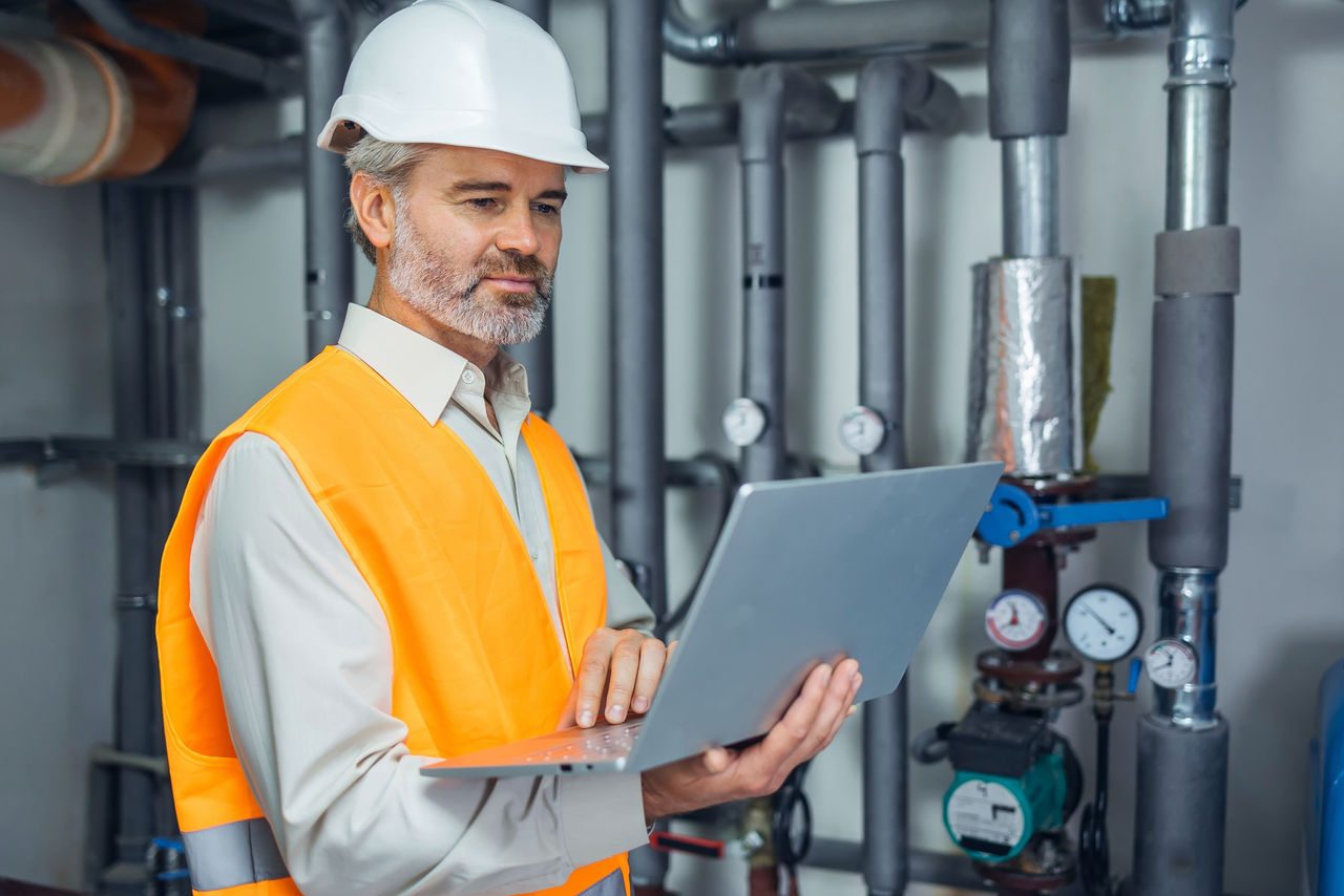 Interno della centrale di teleriscaldamento e tecnico esperto in piedi vicino alla fonte di energia tramite conduttura, utilizzando un computer portatile.