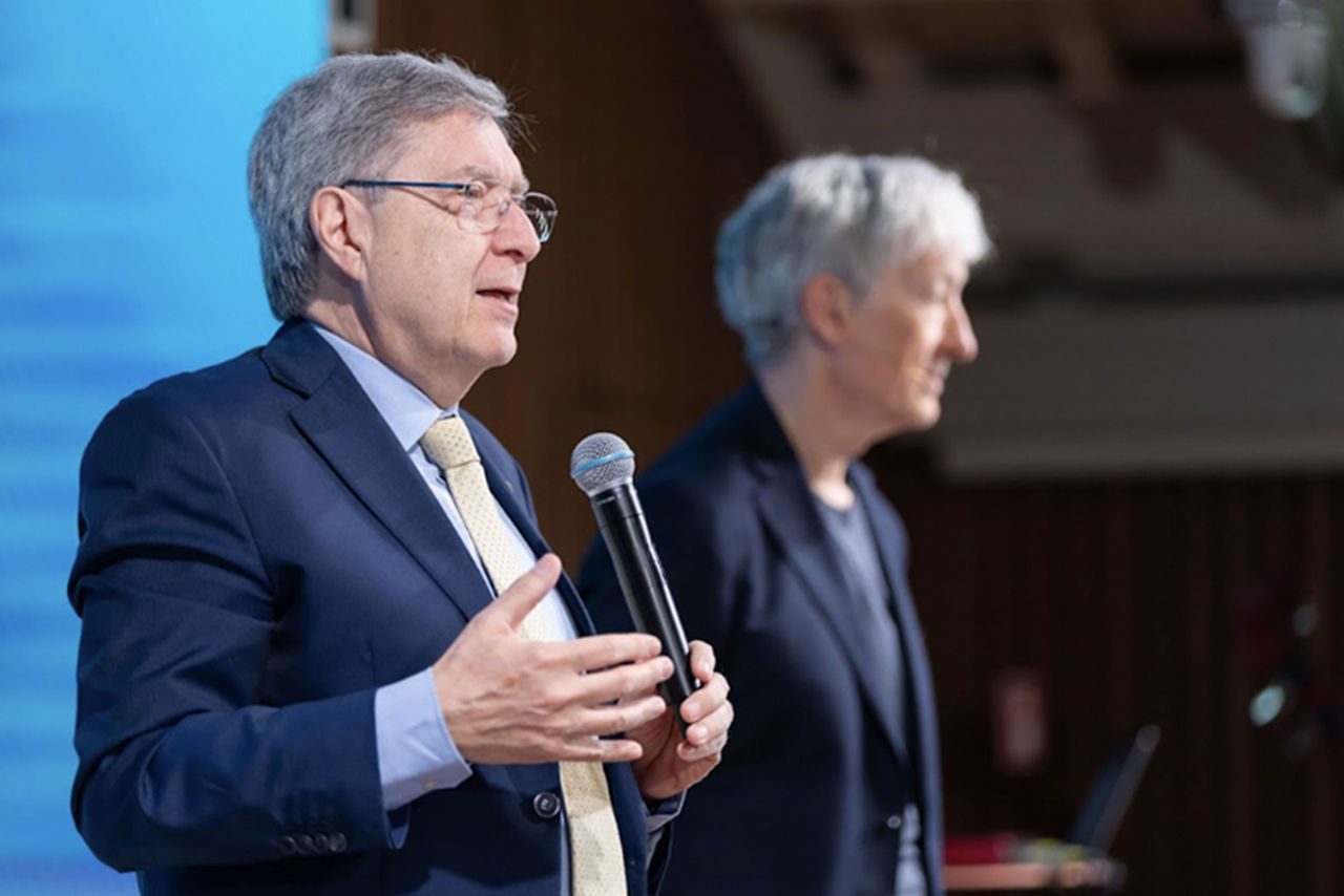 Un relatore in giacca e cravatta parla al microfono durante una conferenza, con un altro speaker sfocato sullo sfondo.