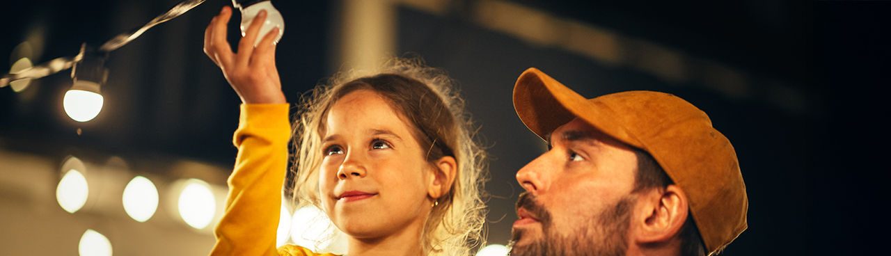 bambina che tocca una lampadina mente si trova in braccio ad un uomo con il berretto 