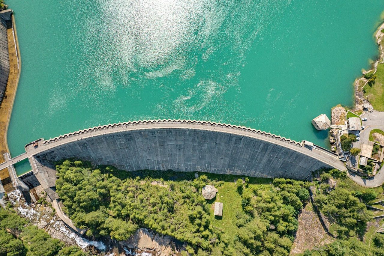 Vista aerea di una diga curvilinea che trattiene le acque di un lago color turchese, circondata da vegetazione e strutture tecniche.