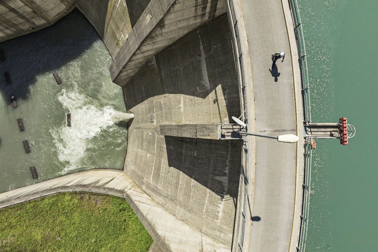 Dettaglio dall'alto di una diga, con getto d'acqua in uscita e persona che cammina sul bordo della struttura, accanto al lago.