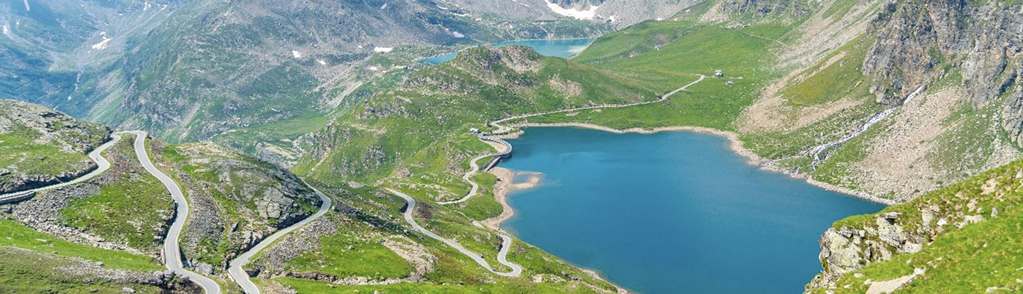 Paesaggio montano con laghi alpini e strade tortuose viste dall'alto, circondato da montagne innevate e prati verdi sotto un cielo azzurro.