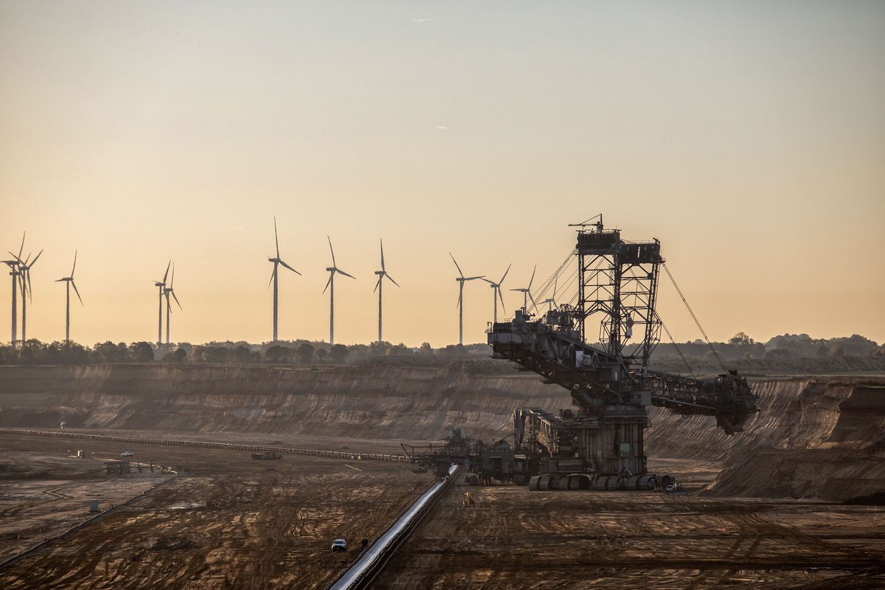 Grande escavatore industriale operante in una miniera a cielo aperto, con turbine eoliche allineate sullo sfondo, rappresentando il contrasto tra le fonti di energia fossile e rinnovabile.
