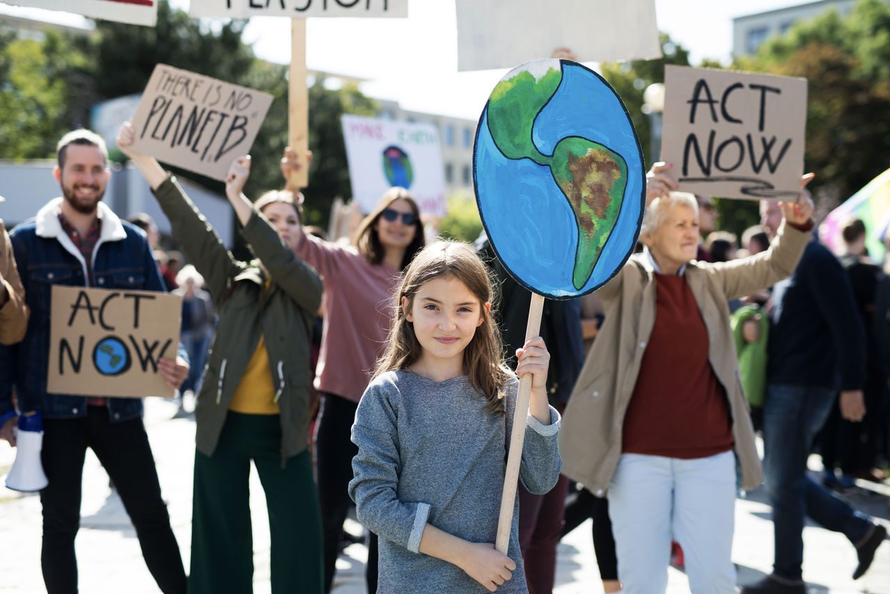 Bambina al centro di una manifestazione per il clima tiene un cartello raffigurante la Terra, circondata da persone con cartelli che recitano slogan come “ACT NOW” e “There is no Planet B”.