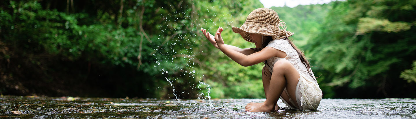 Bambina che gioca seduta in un ruscello, lanciando schizzi d’acqua con le mani, immersa in un ambiente naturale ricco di verde.