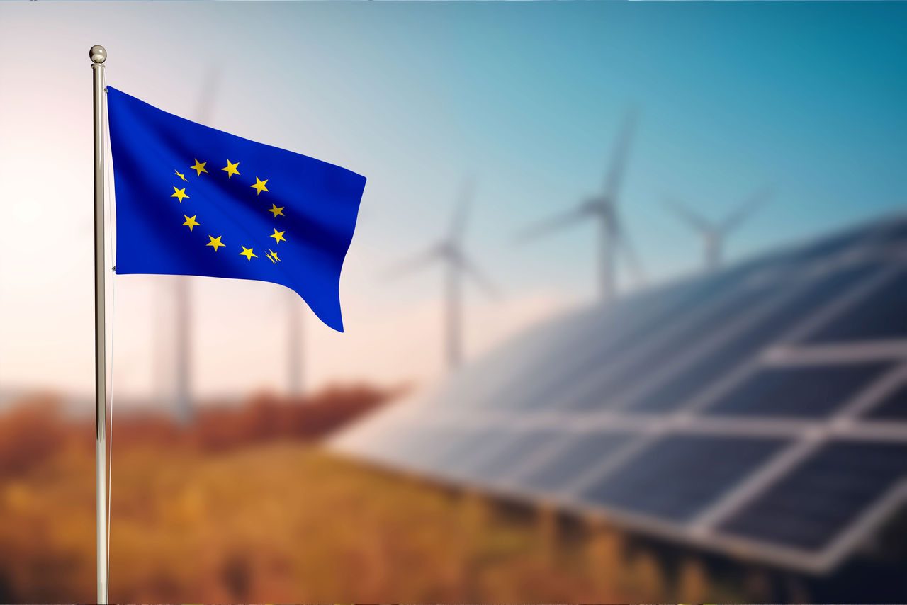 bandiera dell'europa su asta che sventola e come sfondo pannelli fotovoltaici e pale eoliche