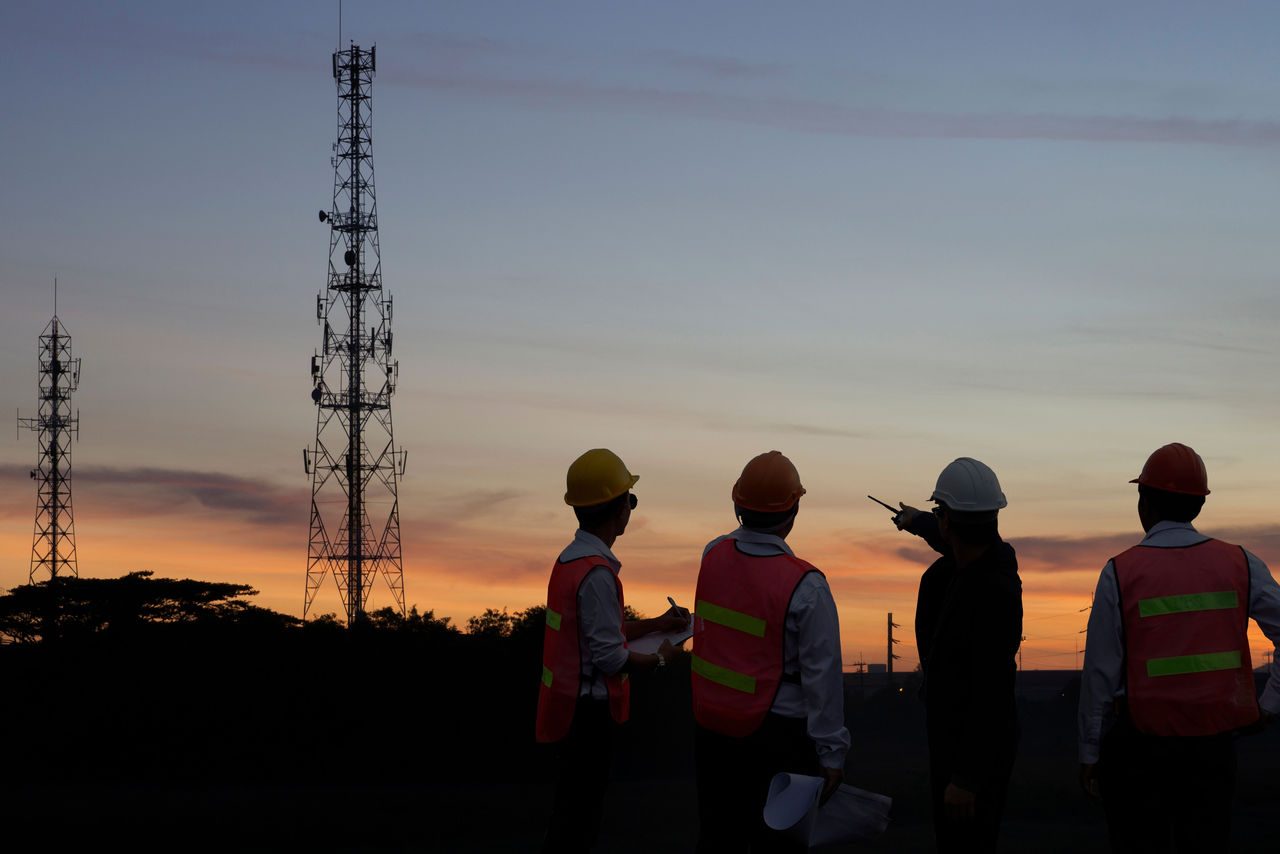 Silhouette di ingegneri e tecnici al tramonto che osservano antenne e strutture per le telecomunicazioni, simbolo dell’innovazione infrastrutturale e del lavoro di squadra.
