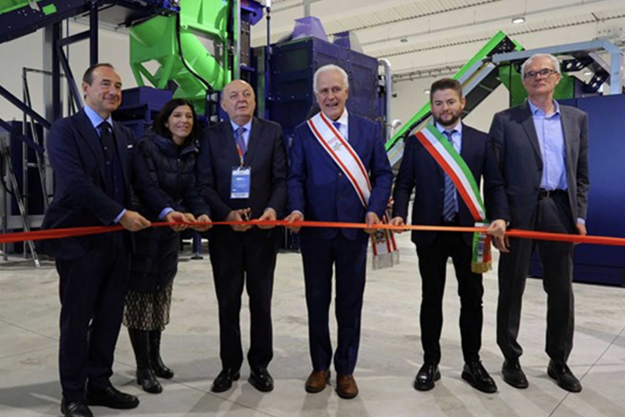 Cerimonia di inaugurazione di un impianto di trattamento rifiuti elettronici, con sei rappresentanti istituzionali che tagliano un nastro rosso davanti a macchinari industriali.