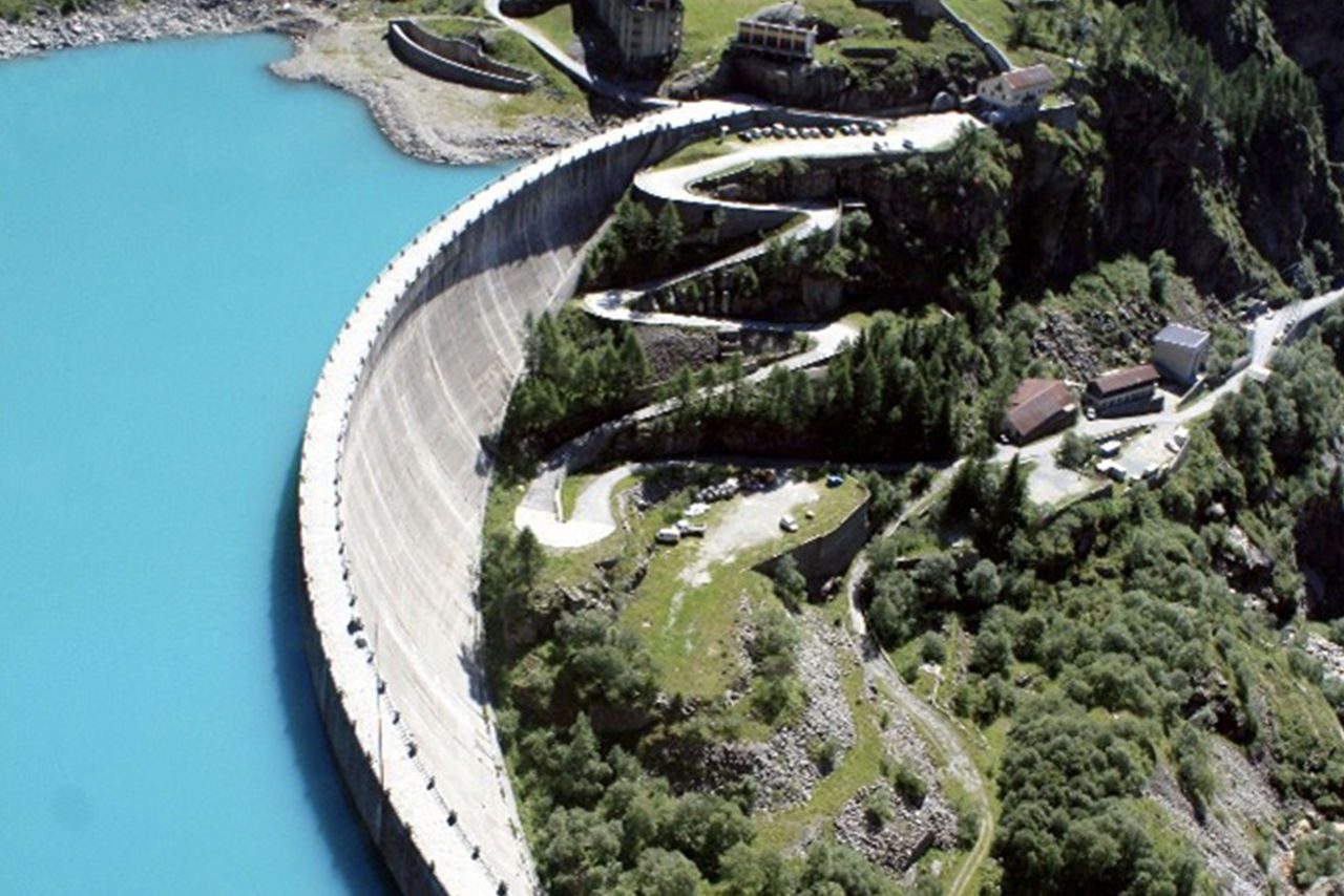 Vista aerea di una grande diga idroelettrica con acqua turchese circondata da un paesaggio montano verde.