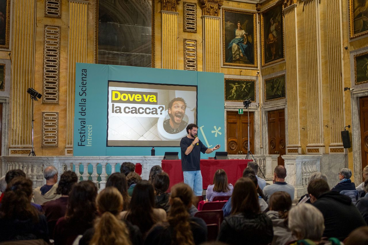 Un divulgatore scientifico di Geopop parla davanti al pubblico al Festival della Scienza di Genova, accanto a un grande schermo con la scritta “Dove va la cacca?”.
