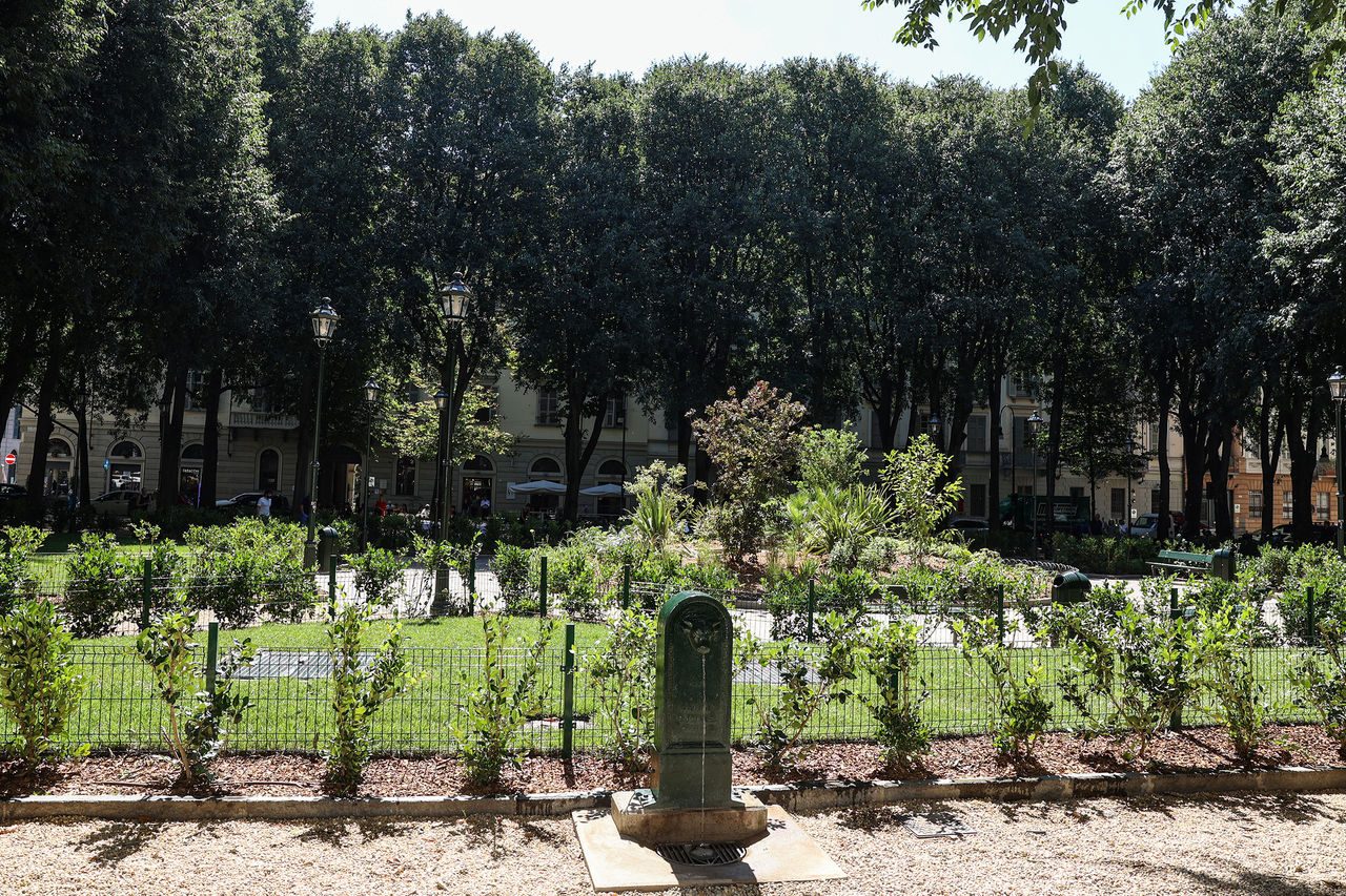Un gruppo di persone, tra adulti e bambini con zainetti blu Iren e cappellini colorati, partecipa a una visita guidata all’aperto, nei pressi di un edificio in pietra e di un campo coltivato.