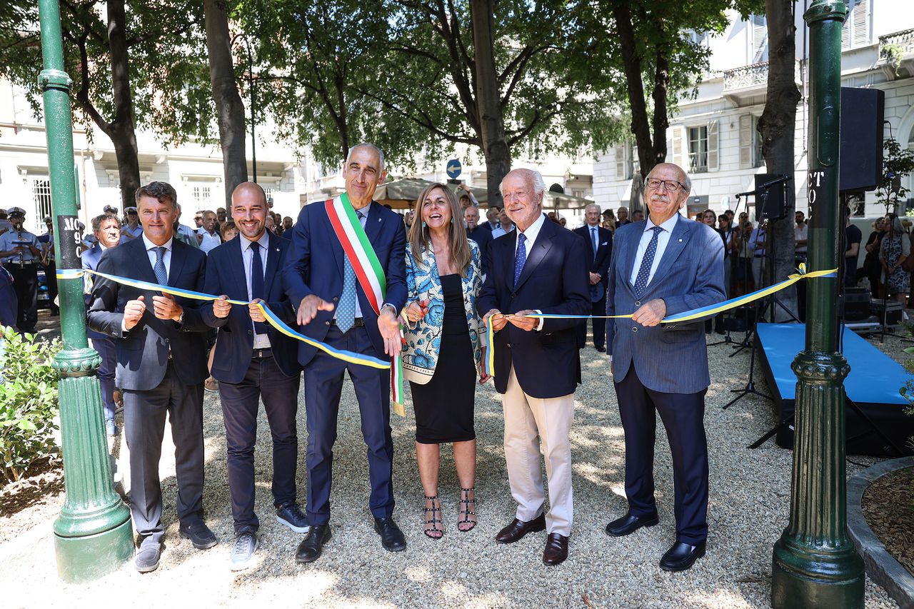 Un gruppo di persone, tra adulti e bambini con zainetti blu Iren e cappellini colorati, partecipa a una visita guidata all’aperto, nei pressi di un edificio in pietra e di un campo coltivato.
