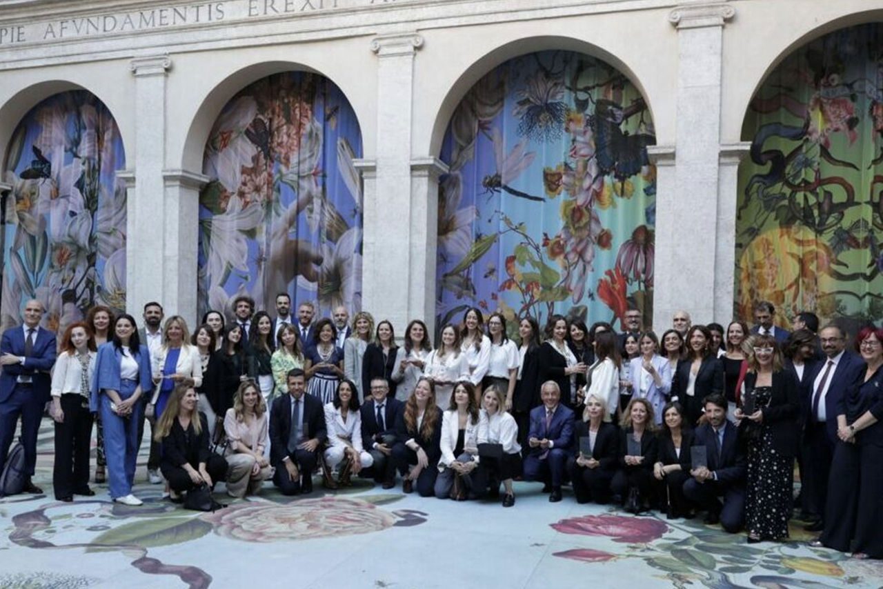 Un ampio gruppo di professionisti e professioniste in abbigliamento formale posa all’interno di un cortile storico con pareti e pavimenti decorati da fiori illustrati. Sullo sfondo, tre grandi archi ornati da drappi floreali.
