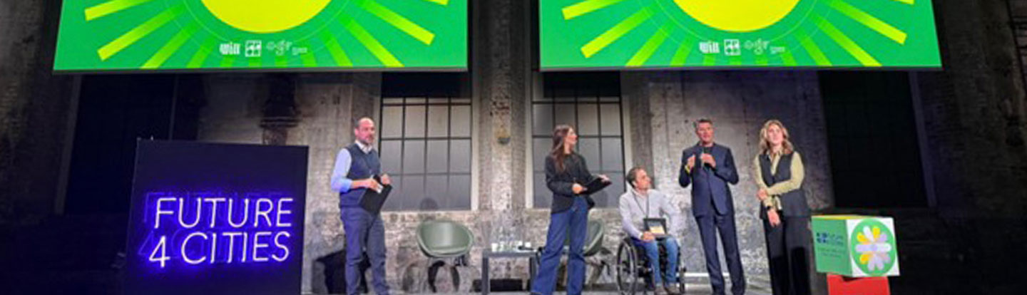 Cinque relatori sul palco durante l’evento Future4Cities, con due schermi verdi sullo sfondo che annunciano il vincitore del premio e un grande cubo decorativo con il logo dell’evento in primo piano.