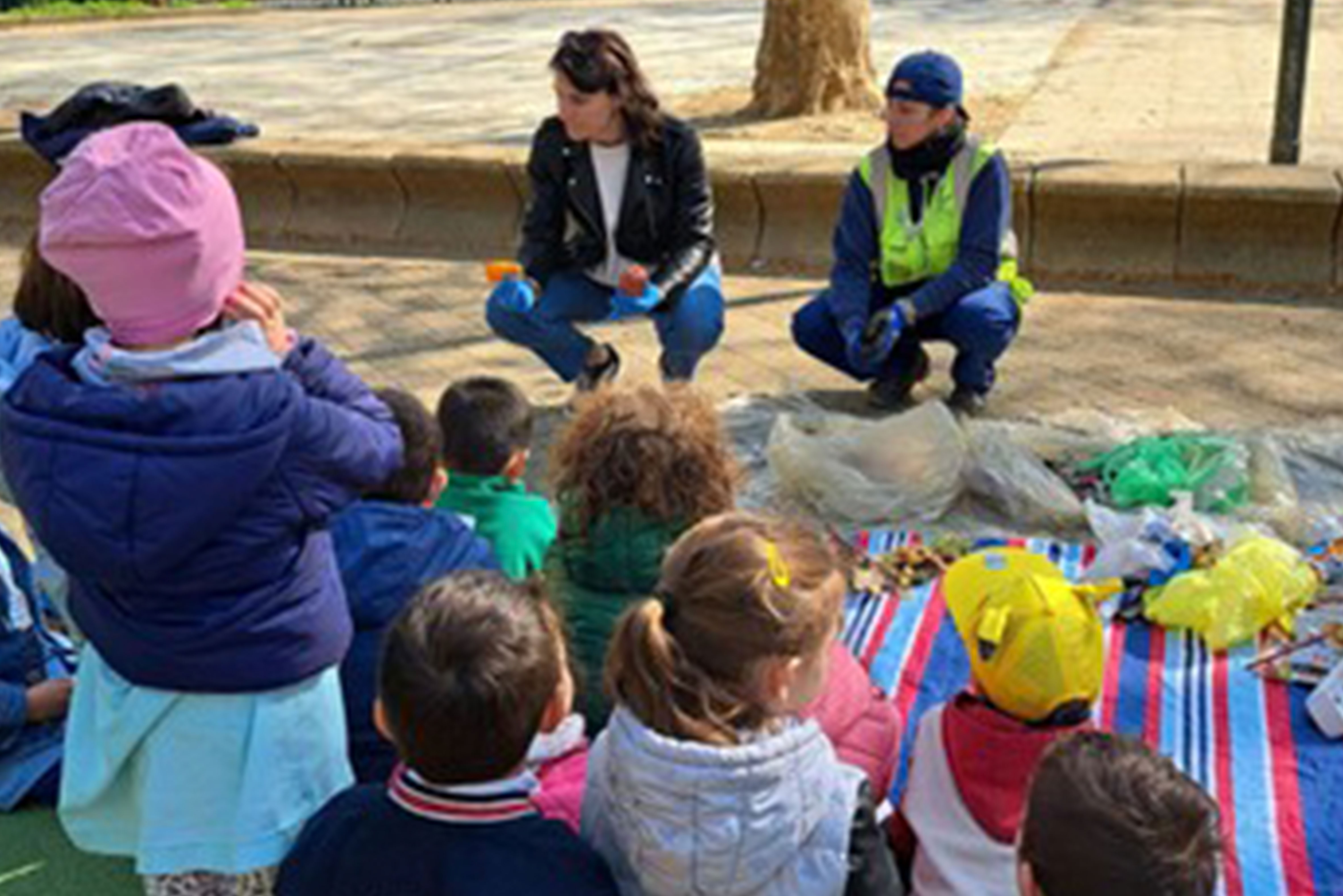 Gruppo di bambini seduti all’aperto ascolta due educatrici che mostrano materiali di scarto su un telo colorato, in un laboratorio ambientale.