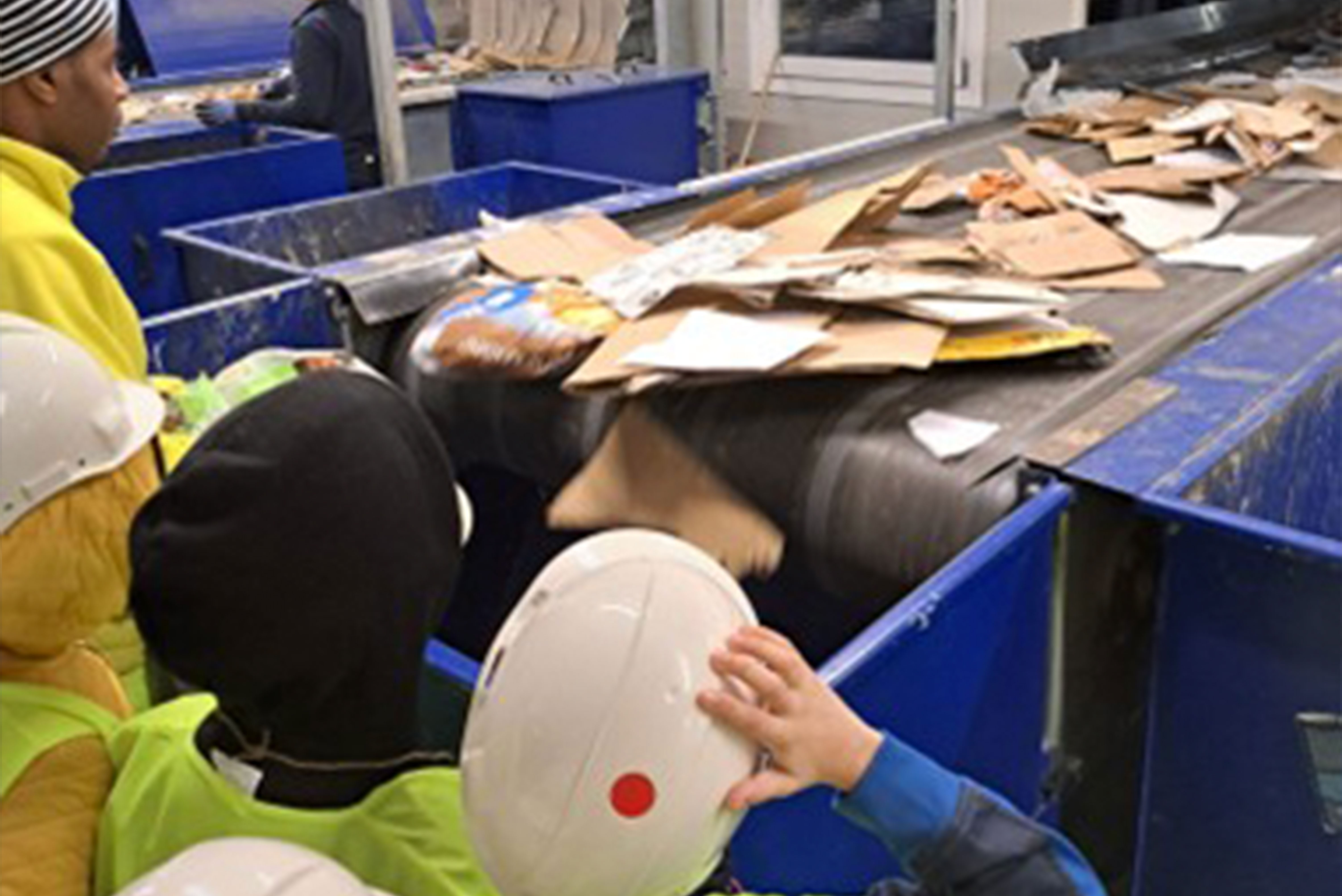 Bambini con caschetti e gilet visitano un impianto di selezione rifiuti, osservando un nastro trasportatore con cartoni da riciclare.