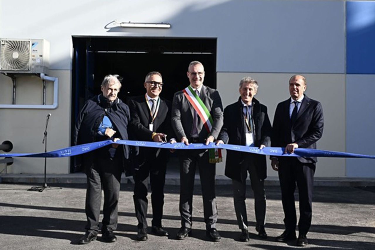 Cerimonia di inaugurazione con cinque uomini in abiti formali che tagliano un nastro blu davanti all’ingresso di un edificio industriale, tra cui un rappresentante con la fascia tricolore.