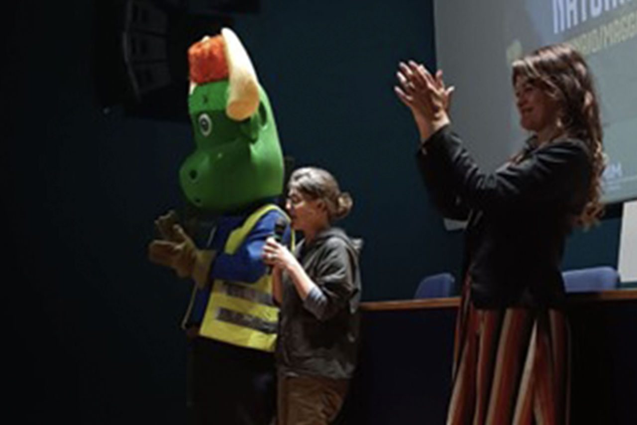 Mascotte verde a forma di drago con giubbotto catarifrangente e donna con microfono sul palco con alle spalle il telo da proiezione di una sala cinematografica, accanto a Paola Bragantini.