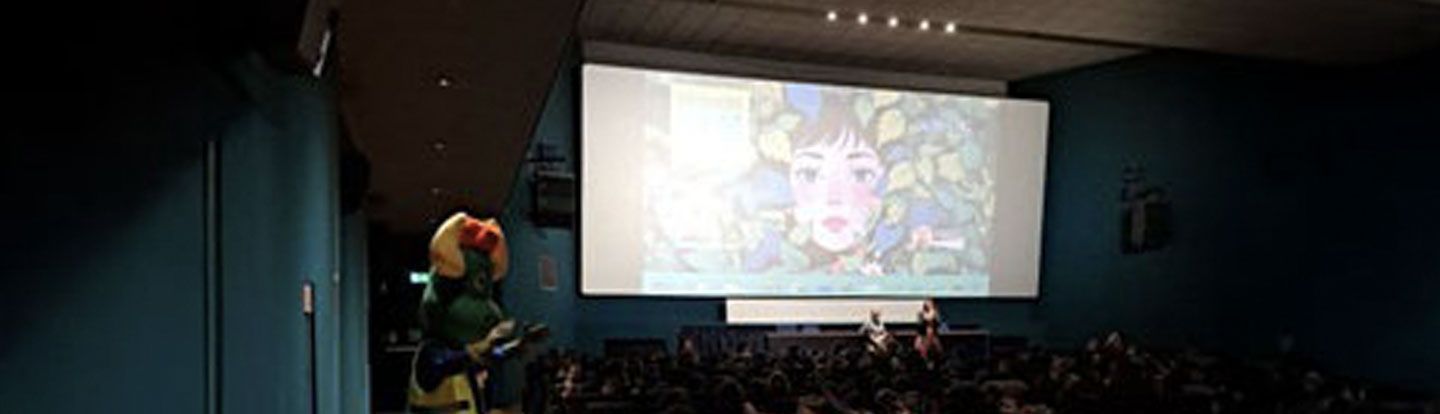 Proiezione in una sala cinematografica con bambini seduti, mascotte verde di Eduiren tra il pubblico, evento educativo dedicato all'ambiente.
