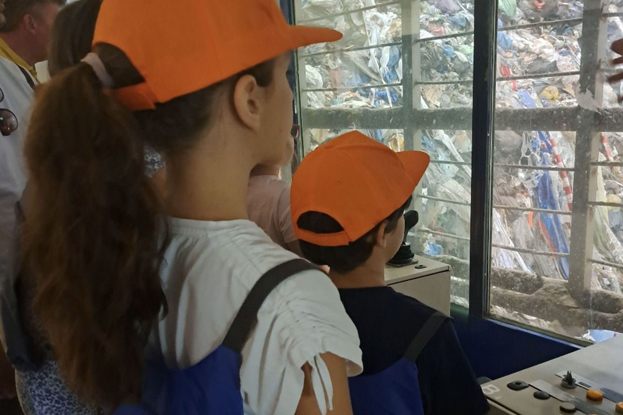 Bambini con cappellini arancioni e zaini Iren osservano un impianto di trattamento rifiuti attraverso una vetrata, durante una visita educativa.