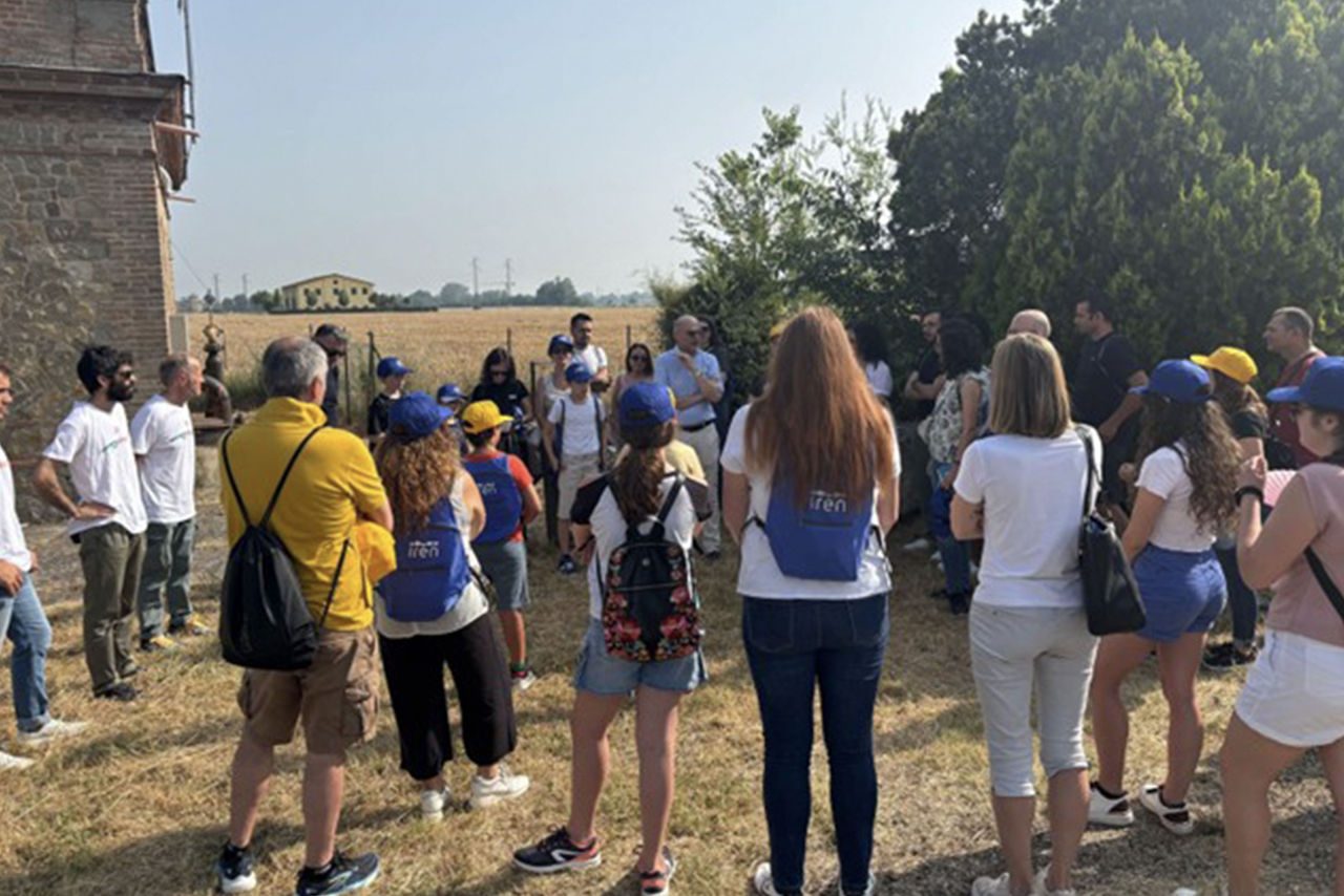 Un gruppo di persone, tra adulti e bambini con zainetti blu Iren e cappellini colorati, partecipa a una visita guidata all’aperto, nei pressi di un edificio in pietra e di un campo coltivato.