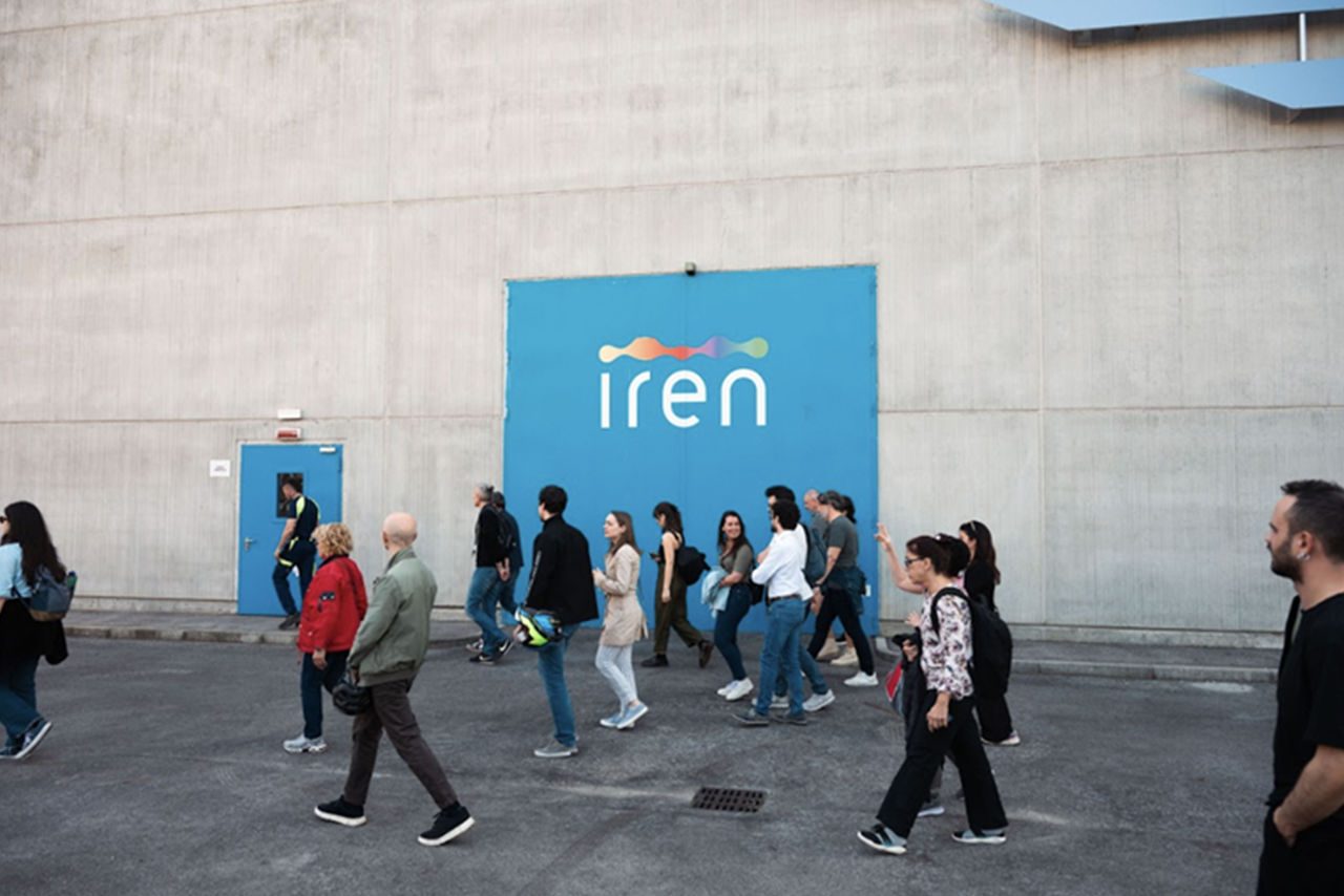 Un gruppo di visitatori entra nel termovalorizzatore Iren, riconoscibile dal grande logo su fondo azzurro posto sul muro esterno dell’edificio.
