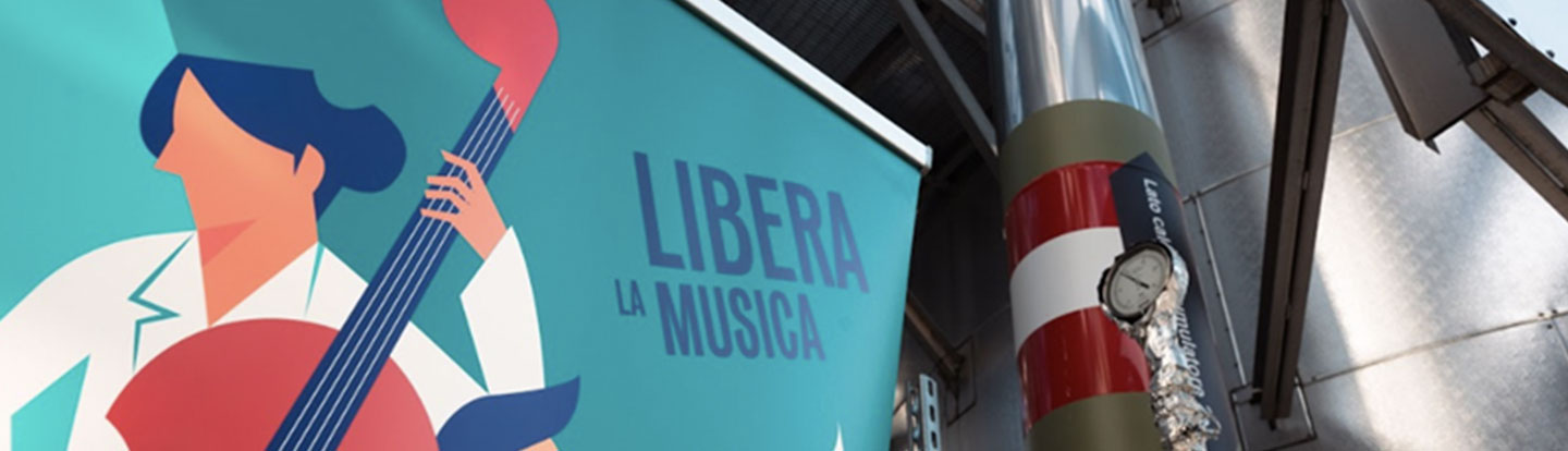 Manifesto illustrato con la scritta “Libera la musica” affisso su una parete del termovalorizzatore Iren, accanto a un impianto industriale decorato per l’evento.