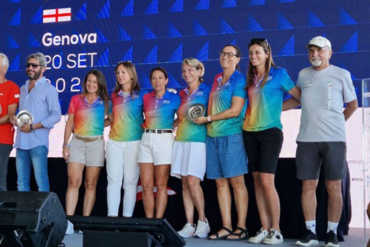 Un gruppo di donne in maglia arcobaleno Iren e pantaloncini estivi posano sorridenti sul palco della Millevele Iren a Genova, durante la premiazione dell’edizione 2025.