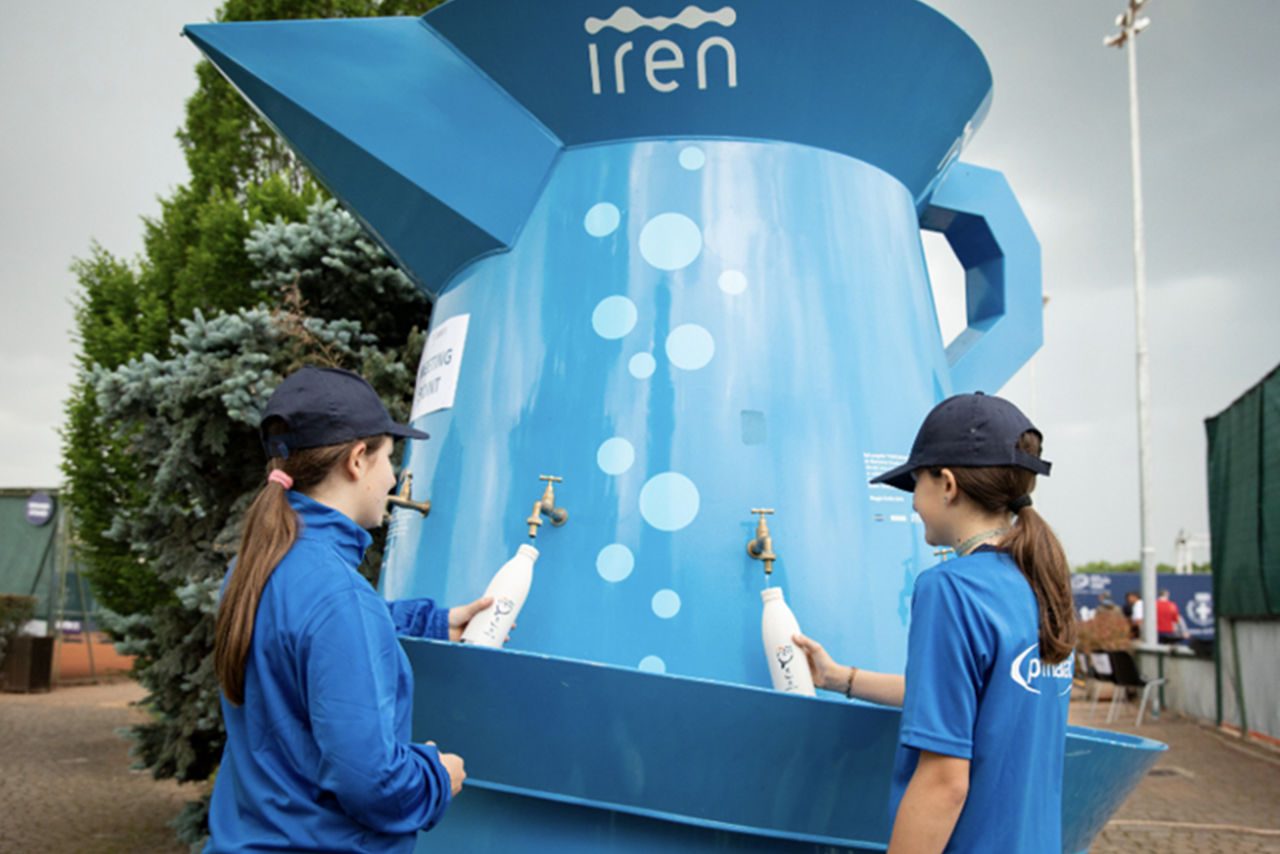 Due ragazze in divisa sportiva riempiono borracce da un grande dispenser d'acqua a forma di caraffa brandizzata Iren.