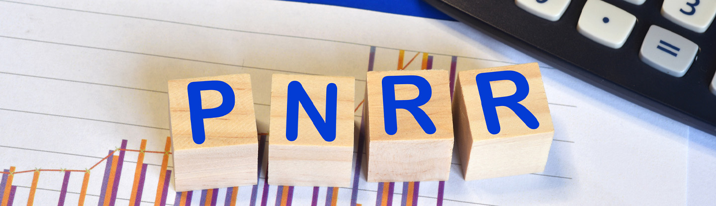 Cubi in legno con le lettere blu "PNRR" poggiati su un foglio con grafici a barre e una calcolatrice, a rappresentare il Piano Nazionale di Ripresa e Resilienza in ambito economico e amministrativo.