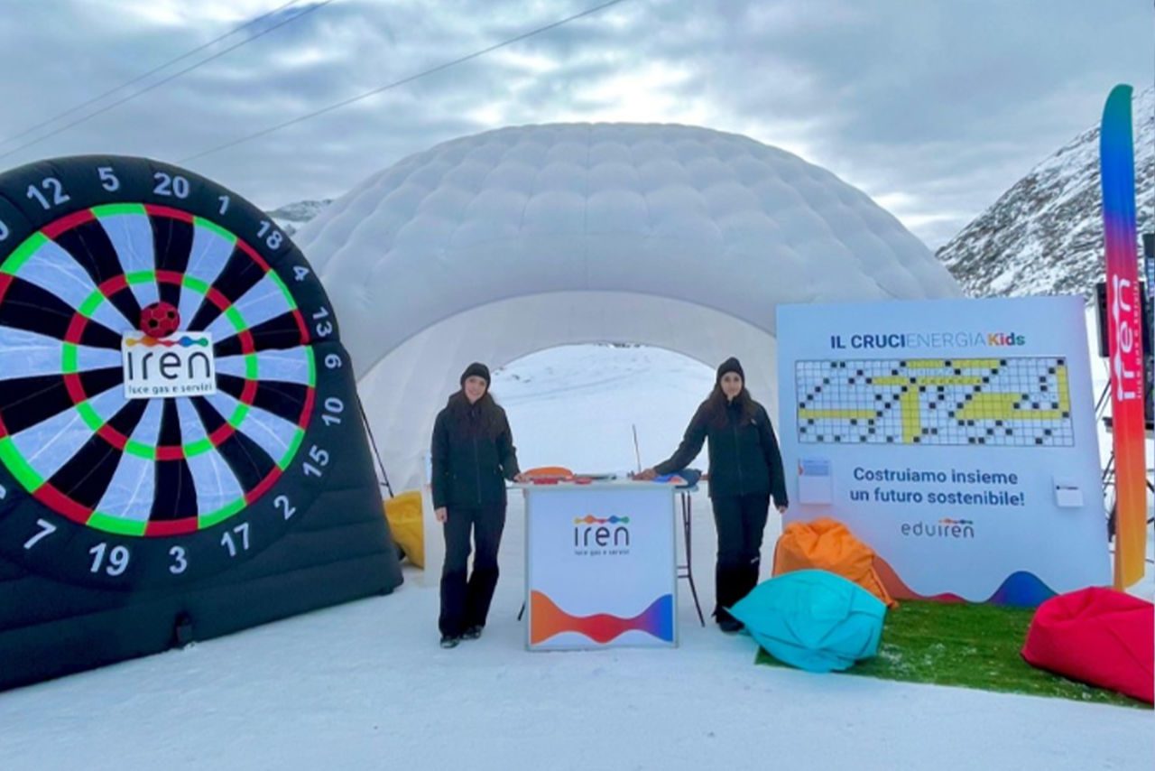 Stand promozionale di Iren su una pista da sci, con un gonfiabile a forma di bersaglio e un cruciverba educativo per bambini, circondato da pouf colorati.