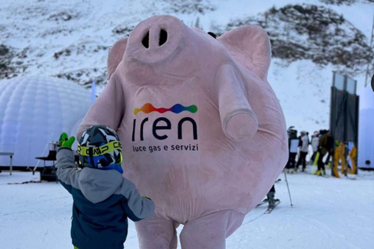 Un bambino con casco da sci interagisce con una mascotte gonfiabile a forma di maialino con il logo Iren, in un contesto di neve e montagne.
