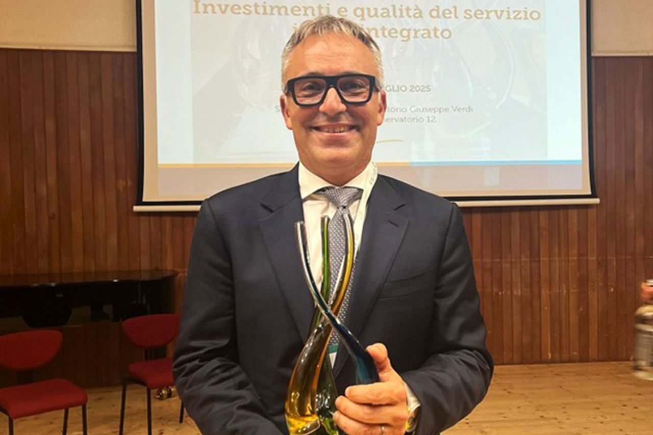 Uomo premiato in giacca e cravatta che sorride tenendo in mano un premio di design in vetro, sullo sfondo una slide di presentazione durante l’evento “Investimenti e qualità del servizio idrico integrato”.