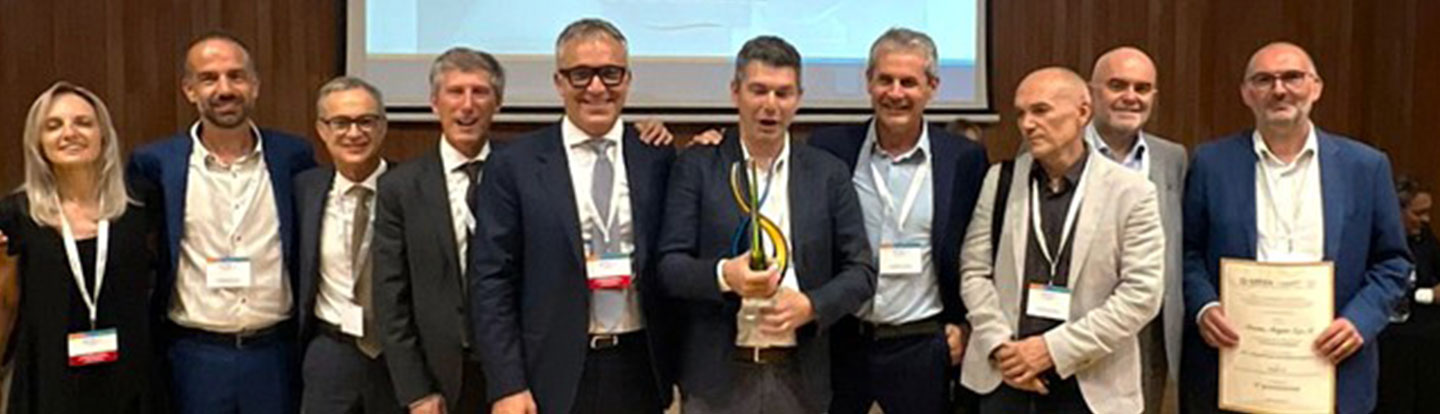Foto di gruppo sul palco con manager e collaboratori premiati con targa e trofeo, davanti alla proiezione di slide dell’evento.