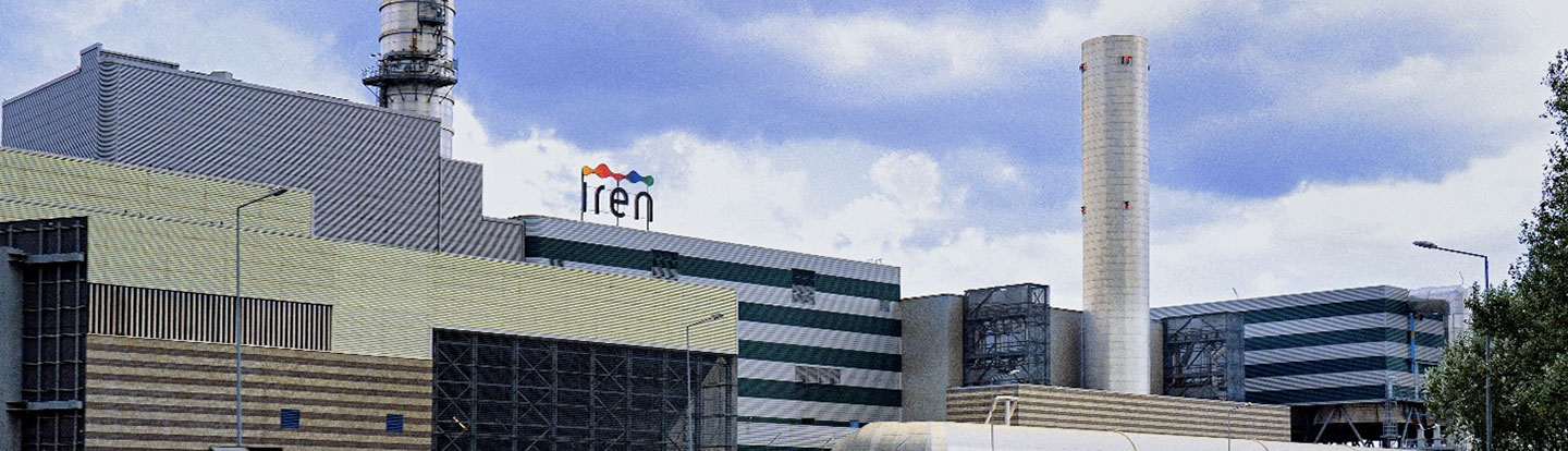 Esterno di un grande impianto Iren con ciminiere e struttura industriale, visibile da una strada a scorrimento veloce.