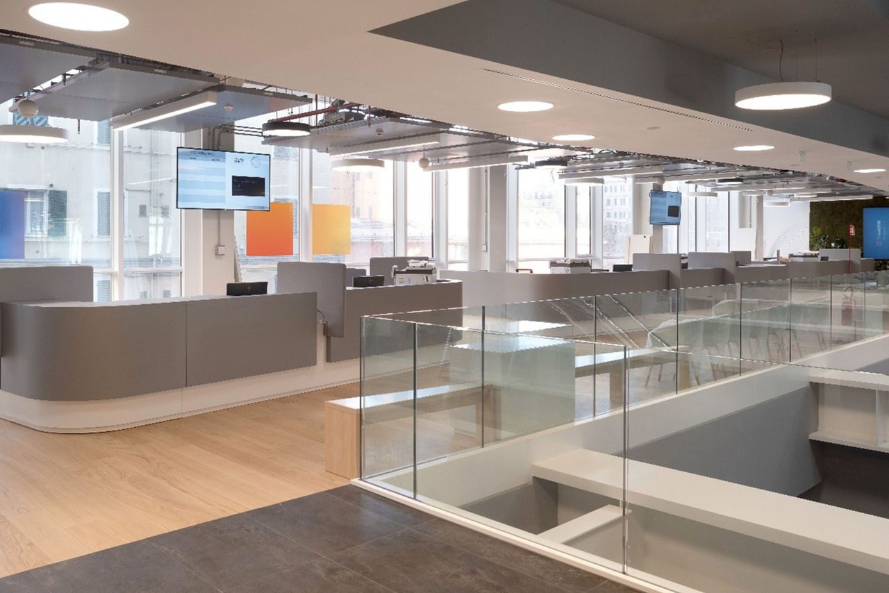 Area reception dello sportello in un ufficio moderno, con postazioni di lavoro, ampie vetrate e un design minimalista con dettagli in vetro e legno.