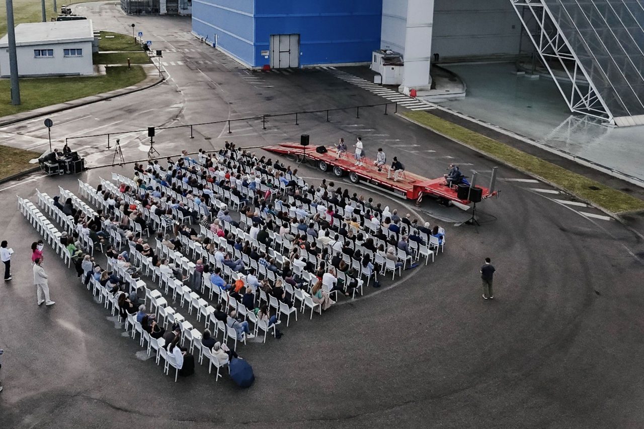 Veduta aerea di un evento all’aperto con un pubblico numeroso seduto su file ordinate di sedie bianche, di fronte a un palco rosso allestito su una piattaforma nel piazzale dell'impianto TRM di Iren.
