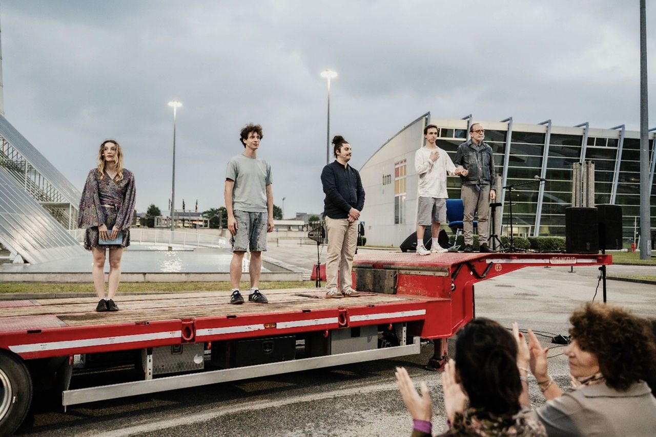 Sei performer in piedi su un palco allestito su un rimorchio rosso, di fronte all’impianto TRM di Torino, durante una performance teatrale all’aperto con microfoni e impianto audio.