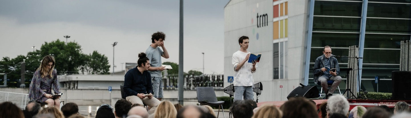 Panoramica laterale del palco su ruote con sei attori in scena durante uno spettacolo teatrale organizzato davanti all’impianto TRM, con pubblico visibile in primo piano.