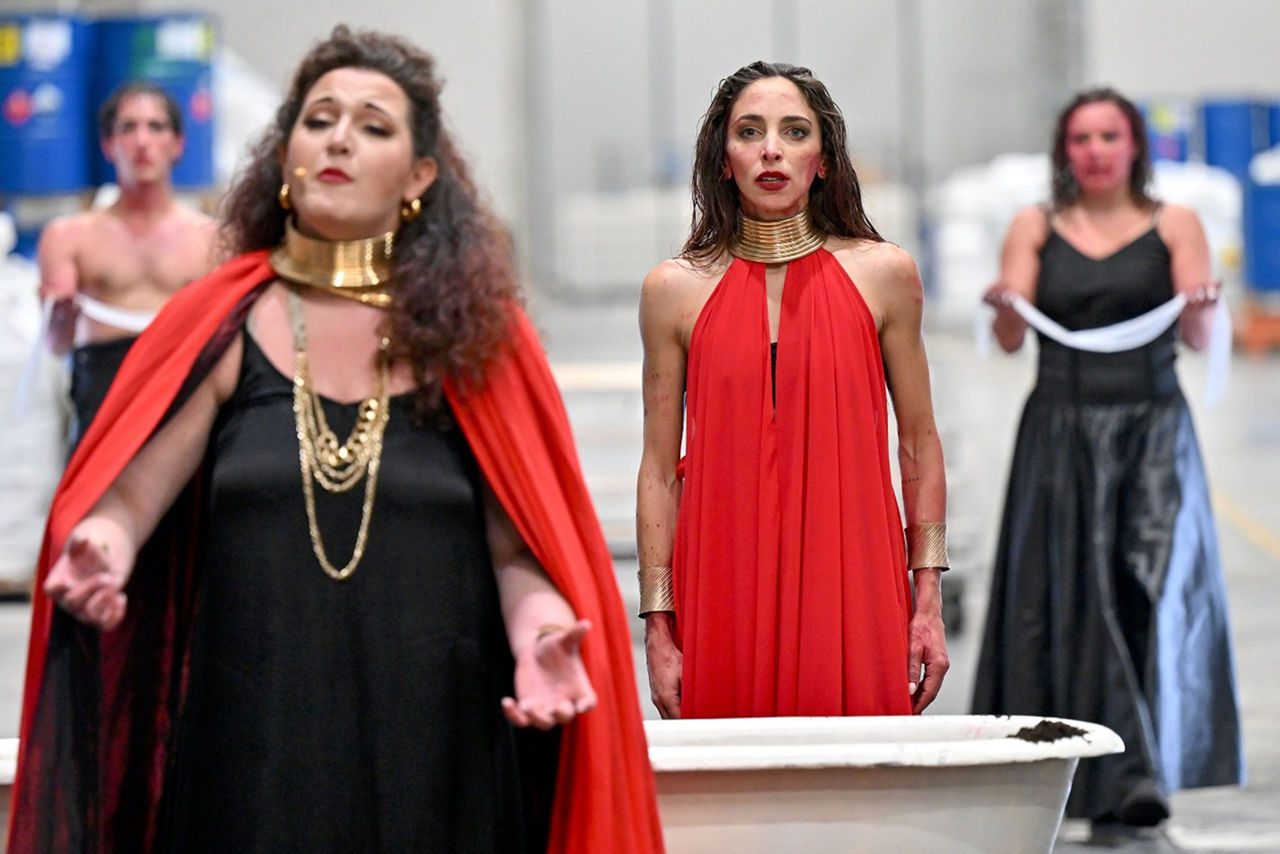 Due interpreti femminili in primo piano, una con mantello rosso e abito nero, l’altra in abito rosso, durante una performance teatrale in un impianto industriale; altri attori sullo sfondo.