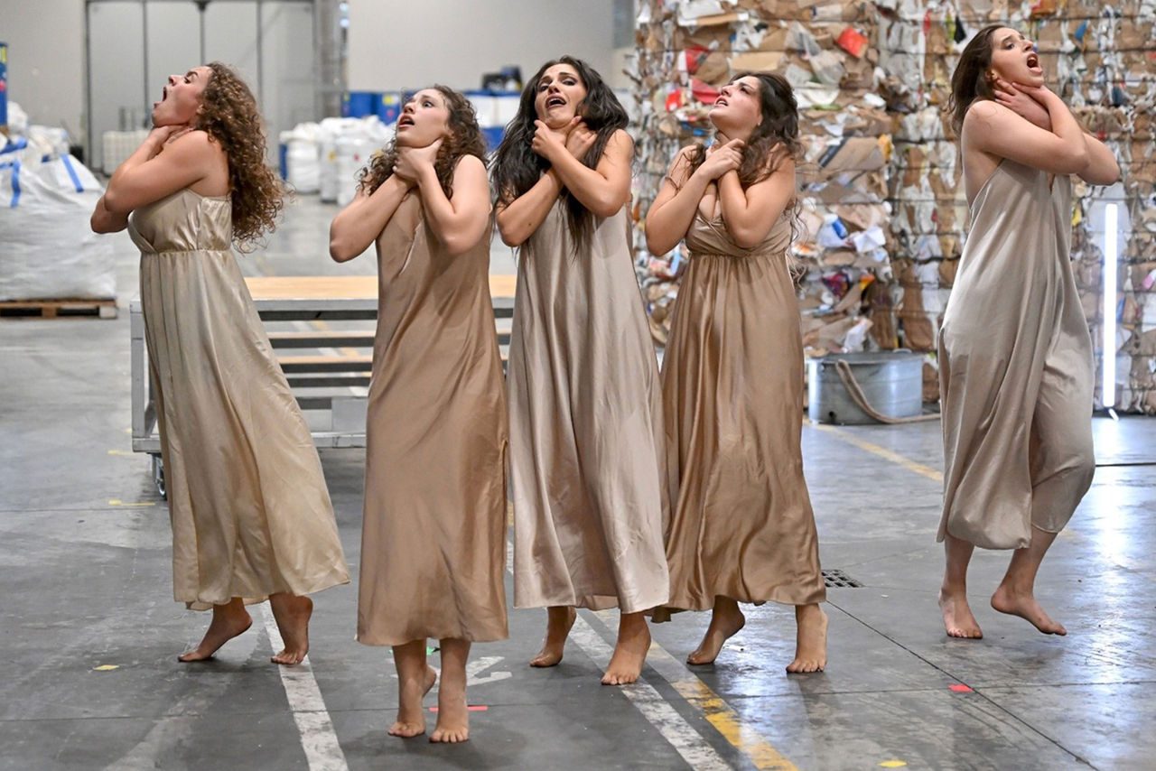 Cinque danzatrici in abiti lunghi color carne si esibiscono in una coreografia intensa all’interno di un capannone industriale, con balle di carta pressata sullo sfondo.