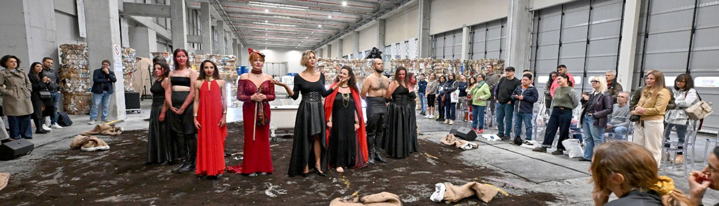 Il cast dello spettacolo in abiti scenici saluta il pubblico al termine della performance, all’interno di uno spazio industriale con balle di carta pressata e spettatori in cerchio.