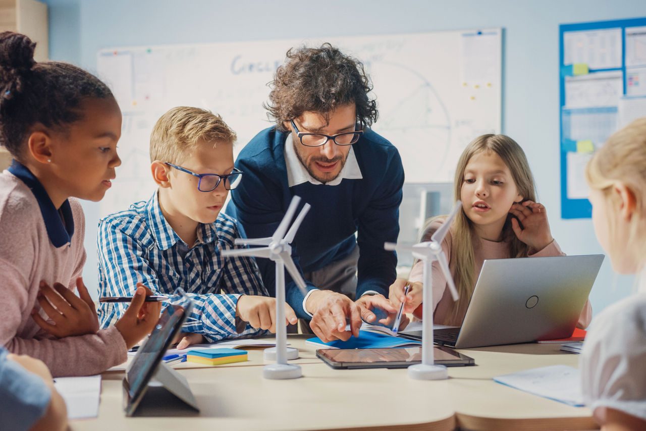 Insegnante e bambini lavorano insieme a un progetto scolastico con modellini di turbine eoliche e tablet, rappresentando l’apprendimento delle energie pulite.