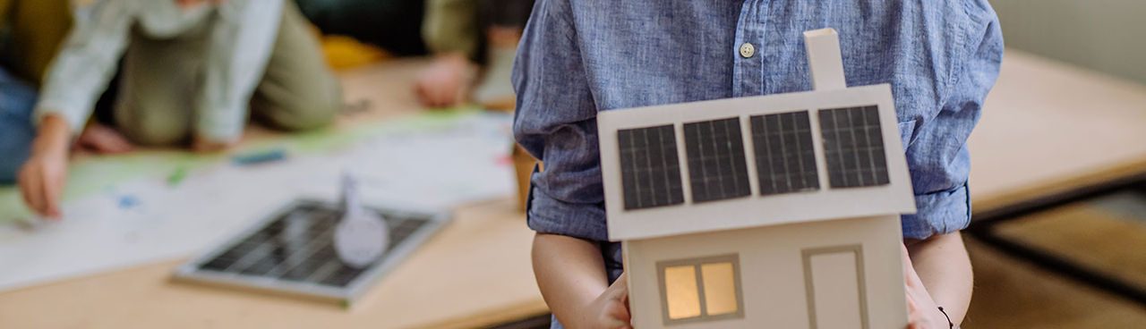Bambino che tiene in mano un modellino di casa con pannelli solari sul tetto, simbolo di educazione alle energie rinnovabili e alla sostenibilità.