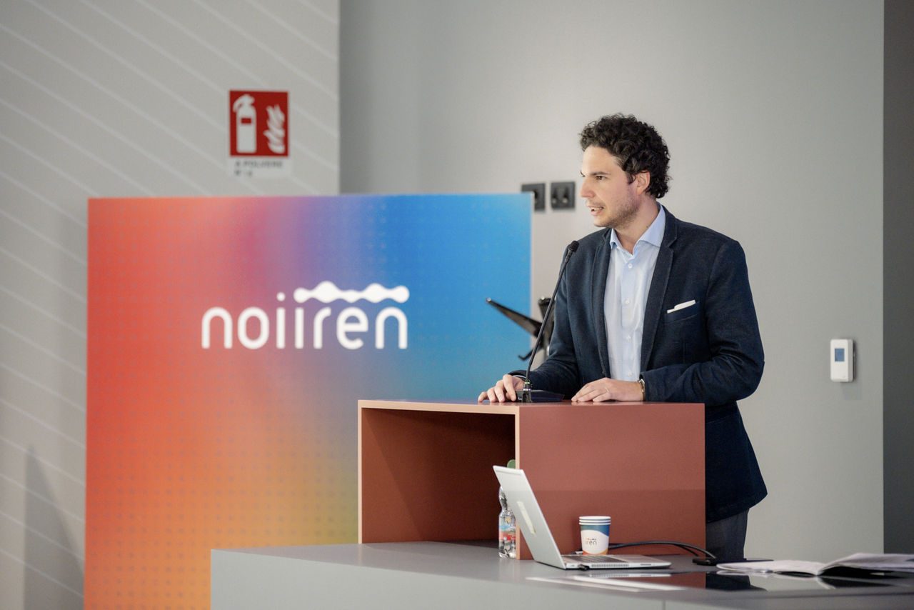 Relatore giovane tiene un discorso dal podio in una sala conferenze Iren, con pannello colorato "noi Iren" sullo sfondo.