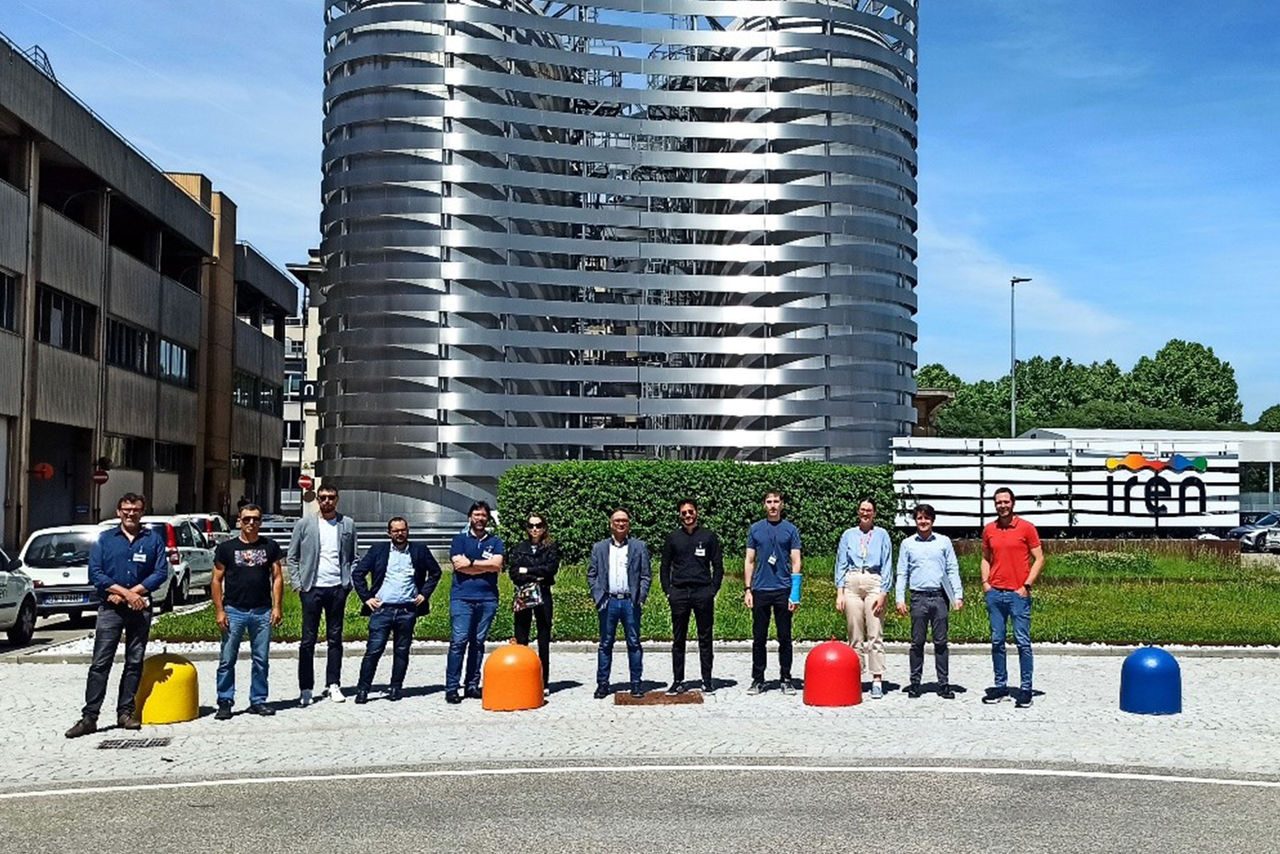 Un gruppo di persone in posa davanti alla sede del Gruppo Iren, con l’edificio in acciaio moderno sullo sfondo e il logo Iren visibile a destra.